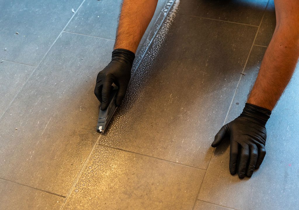 Professional cleaner cleaning grout with a brush blade and foamy soap on a gray tiled bathroom floor